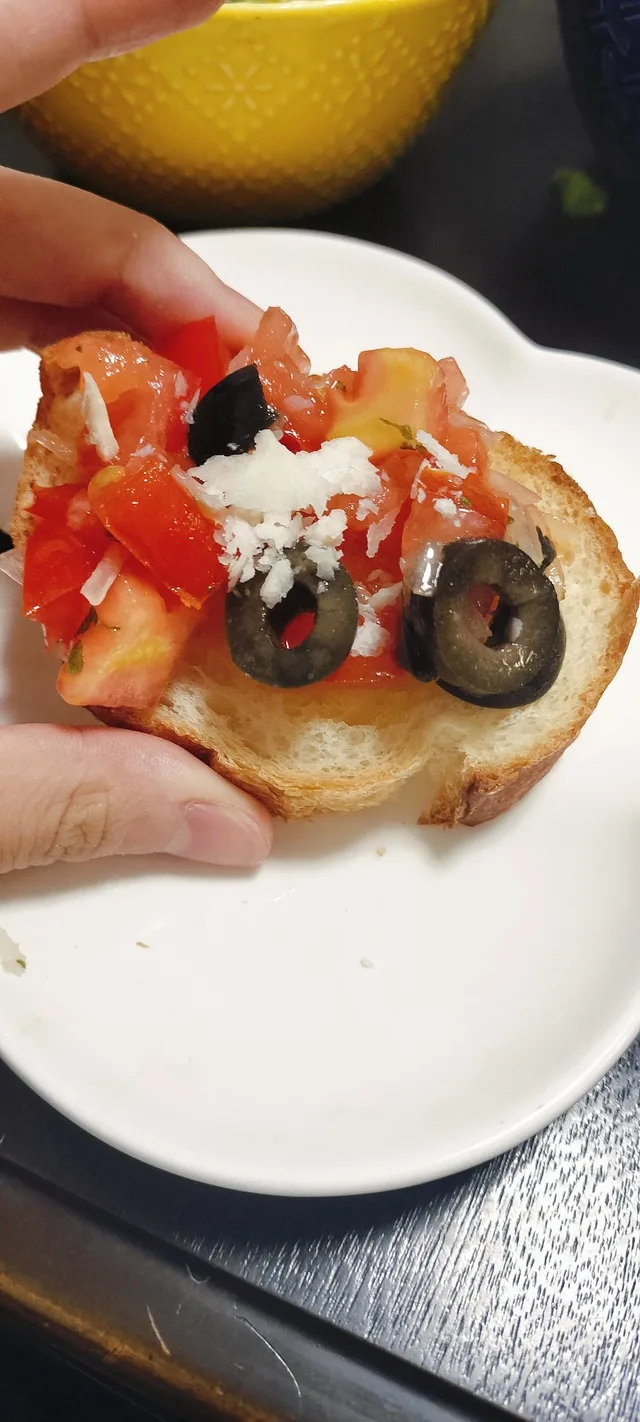 トマトやオリーブ、チーズをのせたパンを手で持っている料理の写真
