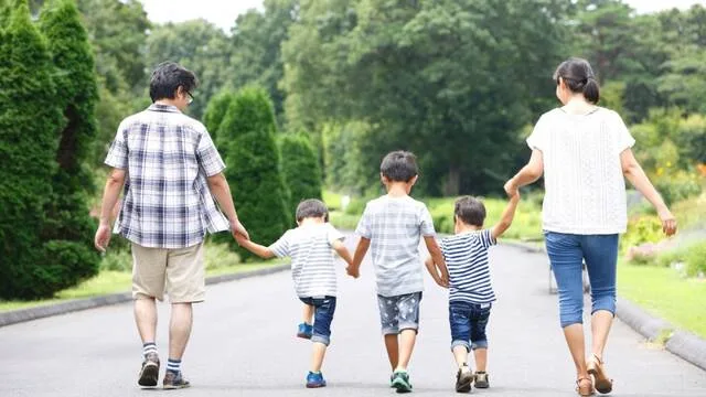両親と3人の子どもが手をつないで緑の多い道を歩いている