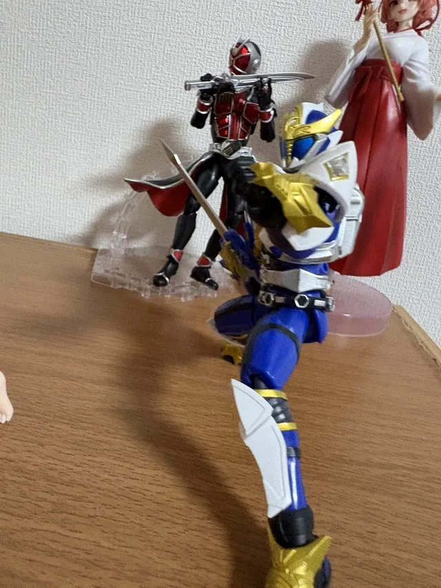 木製の棚に、青と白の仮面ヒーローや黒と赤のフィギュア、赤い衣装の女性フィギュアが飾られている