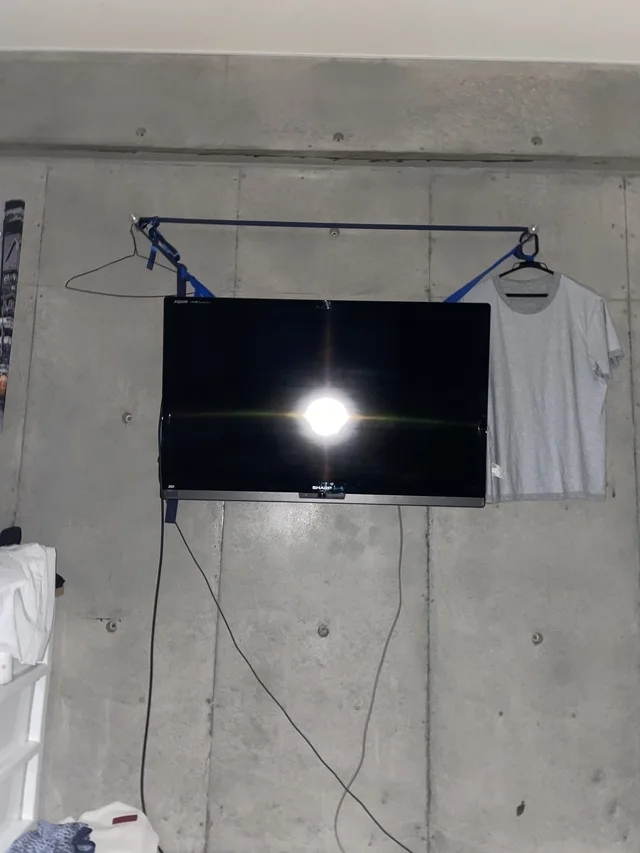 コンクリート壁にベルトで吊るされたテレビと、隣に掛かった白いTシャツ