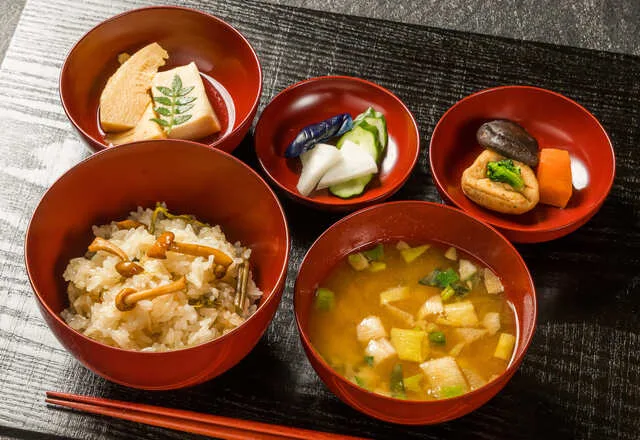 赤い器に盛られた炊き込みご飯、味噌汁、漬物、煮物などの和食膳