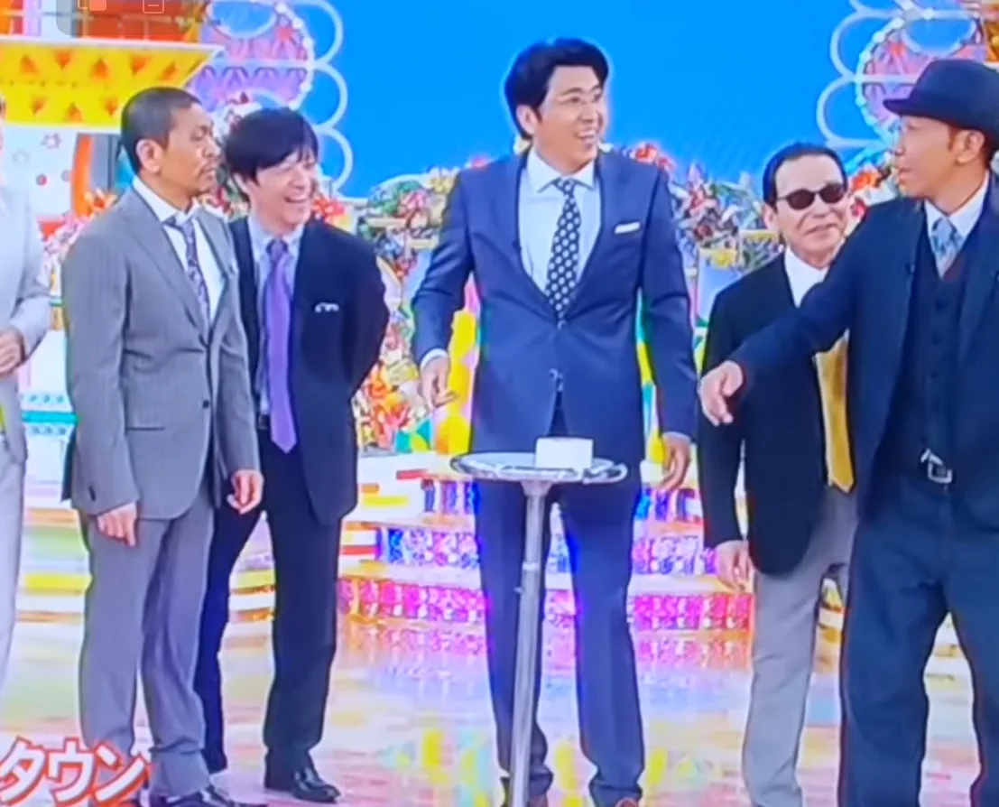 カラフルな舞台で、スーツ姿の男性たちが並んで談笑している