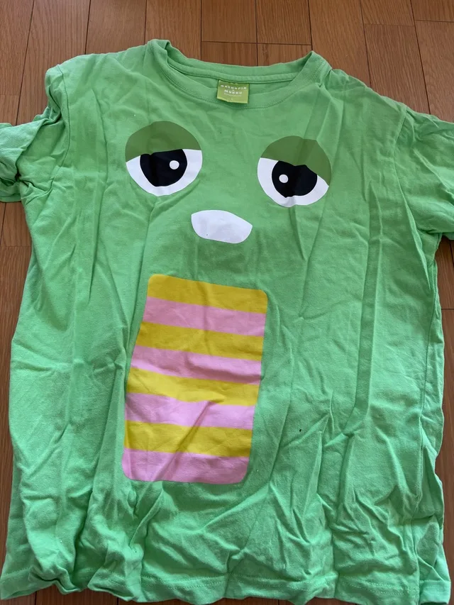 木の床に広げた、目と口、黄色とピンクの縞模様が描かれた緑色のTシャツ。