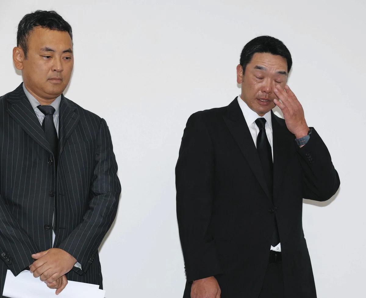 スーツ姿の男性2人が並び、右の男性が目元を拭っている。