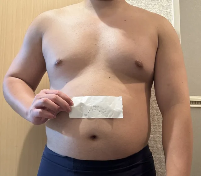 上半身裸の人物が、紙片をお腹の前に持っている。