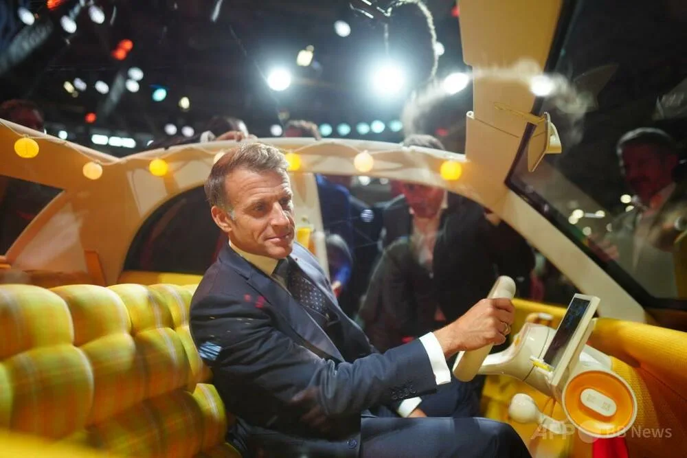 スーツ姿の男性が、照明のある展示会場で車両の運転席に座っている。