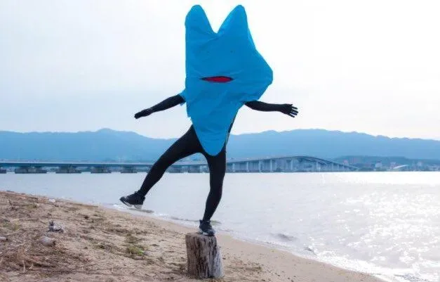 青い着ぐるみ姿の人物が、川辺の杭の上で片足を上げてポーズをしている。
